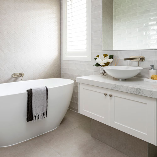 Badrum med gråbeige stengolv, vitt badkar där det hänger en handduk på kanten, vit handfatskommod med vitgå marmorskiva, vitt handfat, kran i guldfärg, spegel och ett fönster med persienner.