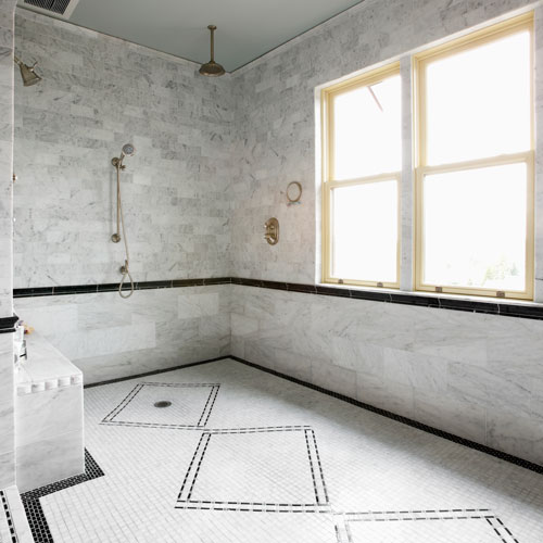Stenlagt ljust mosaikgolv med svart mönster, badrummet har sljust kaklade väggar, stort fönster, dusch och krokar i guldfärg